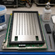 Antminer S21 Hydro hashboard on a repair bench with stainless-steel thermal grease stencil mounted on its positioning frame, 8 W/mK blue silicone gel pail and dispenser gun ready for application — S21 series thermal paste guide